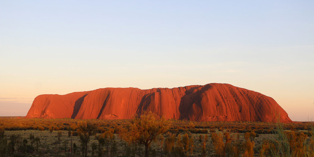 a large red rock in the desert with Uluru in the background