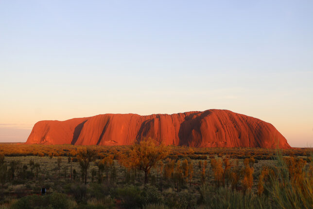 a large red rock in the desert with Uluru in the background