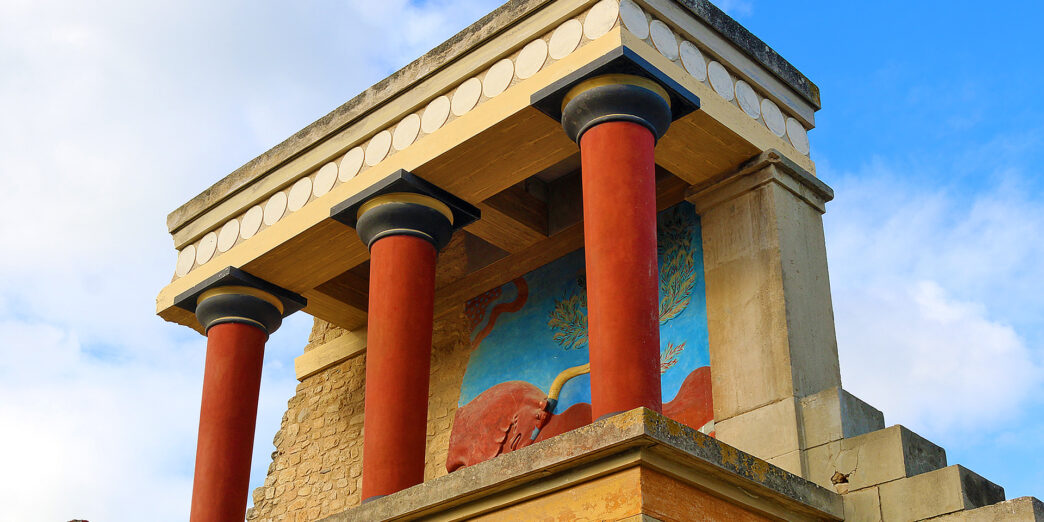 a stone building with columns and a mural on the wall with Knossos in the background