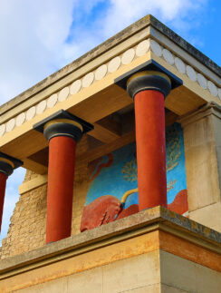 a stone building with columns and a mural on the wall with Knossos in the background