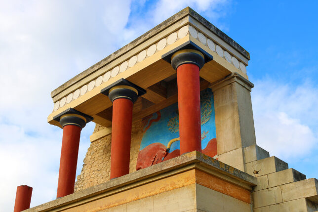 a stone building with columns and a mural on the wall with Knossos in the background