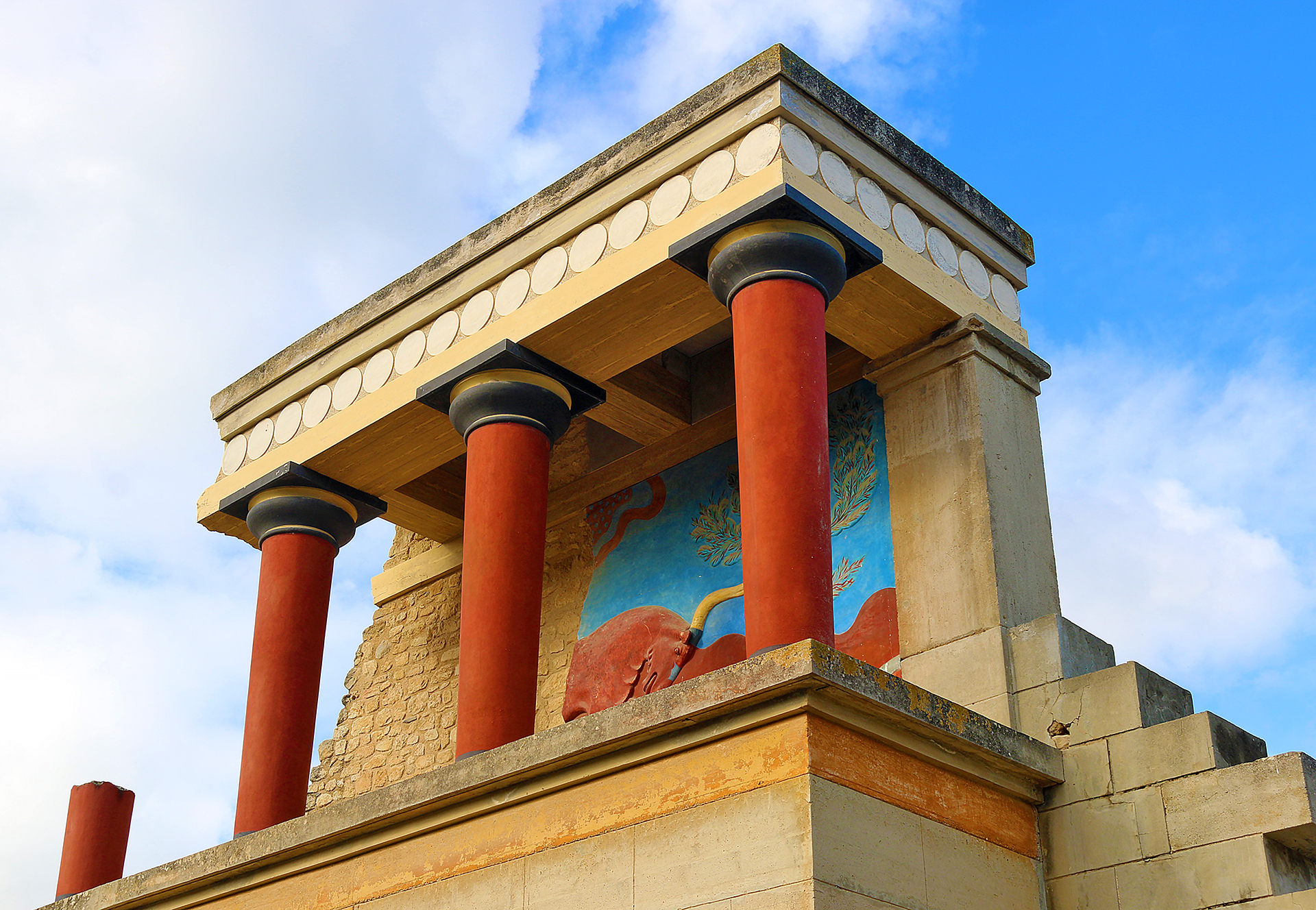 a stone building with columns and a mural on the wall with Knossos in the background