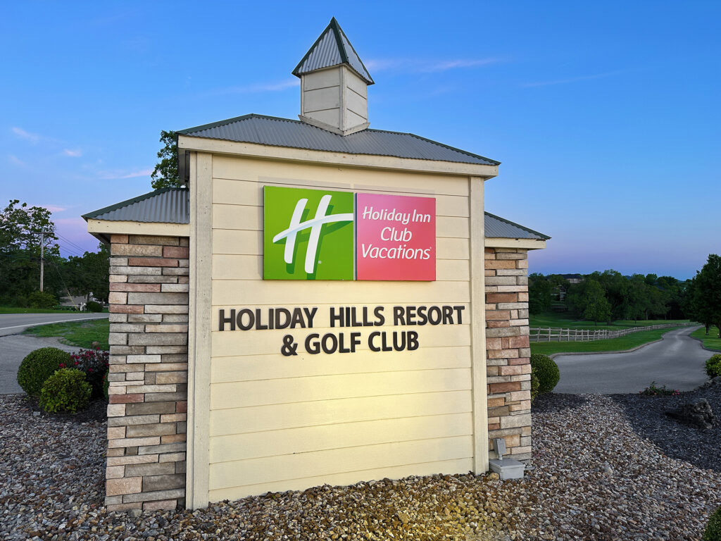 The image shows a sign for "Holiday Inn Club Vacations" at "Holiday Hills Resort & Golf Club." The sign is mounted on a structure with a stone and wood design, featuring a small roof. The background includes a road, greenery, and a clear blue sky.